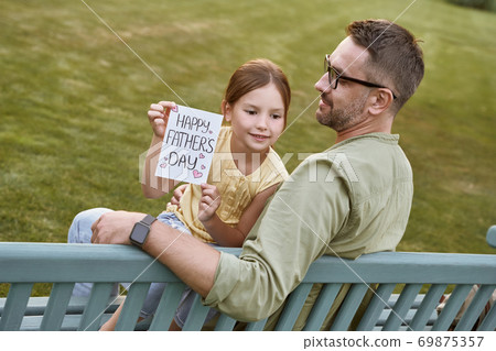 Celebrating Happy Fathers Day outdoors. Young loving father sitting on the wooden bench in park with his cute little daughter, happy girl holding handmade postcard for daddy Celebrating Happy Fathers Day outdoors. Young loving father sitting on the wooden bench in park with his cute little daughter, happy girl holding handmade postcard for daddy 69875357