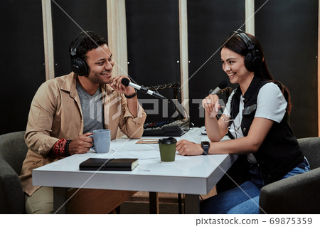 Portait of two happy radio hosts, man and woman smiling while talking with each other, moderating a live show in studio Portait of two happy radio hosts, man and woman smiling while talking with each other, moderating a live show in studio 69875359