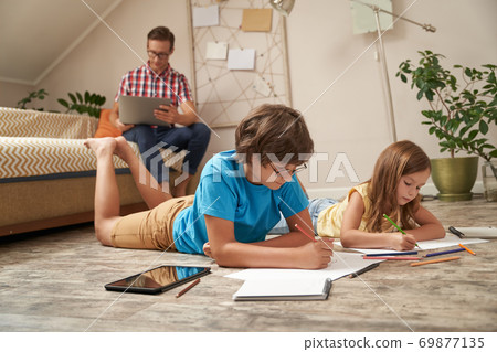 Cute little boy and girl lying on the wooden floor and drawing with colorful pencils while their father sitting on sofa and working on laptop 69877135
