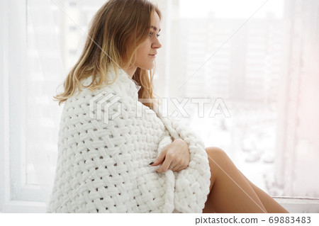 Woman with a white plaid on her shoulders is sitting on the windowsill Woman with a white plaid on her shoulders is sitting on the windowsill 69883483