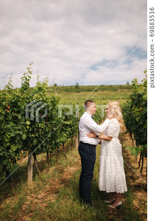 Groom and bride walking in vineyard in their wedding day. Blond hair woman in white dress hold wedding bouquet in hands 69883516