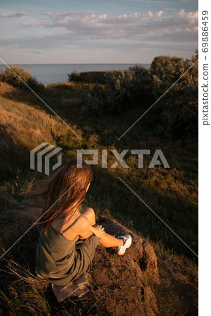 View from the back of a girl in a shirt sitting on a hill and admiring the beautiful view and sunrise or sunset View from the back of a girl in a shirt sitting on a hill and admiring the beautiful view and sunrise or sunset 69886459
