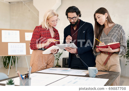 Two businesswomen making notes and looking at display of tablet held by man Two businesswomen making notes and looking at display of tablet held by man 69887708