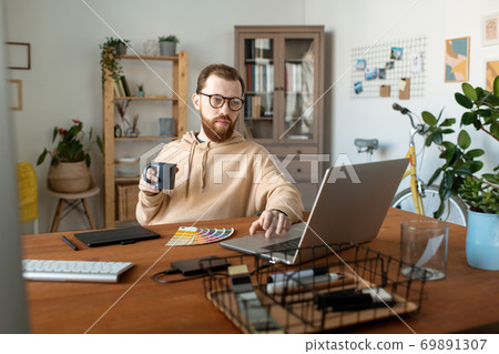 Young bearded freelancer looking at laptop display while having coffee by table 69891307