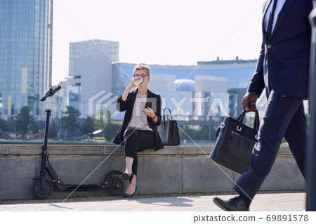 Young relaxed businesswoman scrolling in smartphone and drinking coffee Young relaxed businesswoman scrolling in smartphone and drinking coffee 69891578