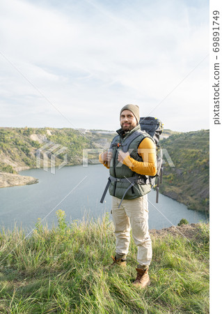 Happy young bearded tourist with backpack standing on top of mountain Happy young bearded tourist with backpack standing on top of mountain 69891749