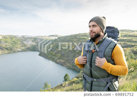 Happy young bearded backpacker moving along mountain against lake or river Happy young bearded backpacker moving along mountain against lake or river 69891752