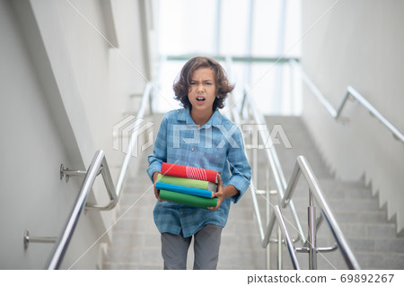 Upset schoolboy walking downstairs, carrying pile of books Upset schoolboy walking downstairs, carrying pile of books 69892267