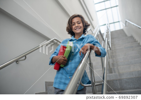 Schoolboy leaning on stair railing, holding pile of books Schoolboy leaning on stair railing, holding pile of books 69892307