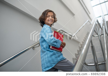Happy schoolboy holding pile of books, walking upstairs Happy schoolboy holding pile of books, walking upstairs 69892308