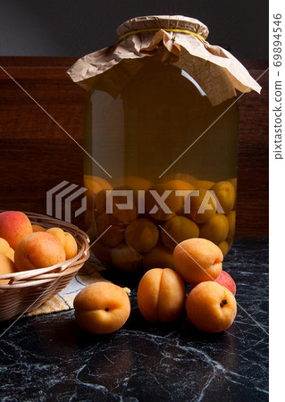 Fresh organic harvested apricots in basket and big glass jar with canned apricot compote on dark marble.. Fresh organic harvested apricots in basket and big glass jar with canned apricot compote on dark marble.. 69894546