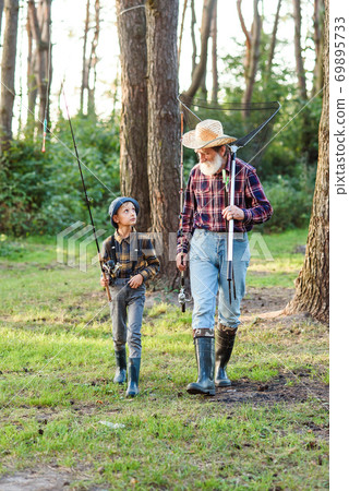 Likable confident bearded grandpa in forest going fishing together with his 10-aged grandson and talking about fishing. Likable confident bearded grandpa in forest going fishing together with his 10-aged grandson and talking about fishing. 69895733