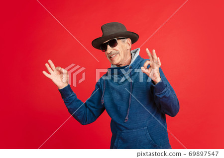 Senior man in stylish eyewear and hat isolated on red background. Tech and joyful elderly lifestyle concept Senior man in stylish eyewear and hat isolated on red background. Tech and joyful elderly lifestyle concept 69897547