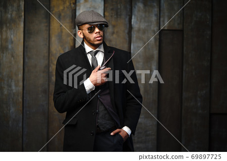 Studio shot of stylish african american man wearing coat, cap, glasses and holding smoking pipe Studio shot of stylish african american man wearing coat, cap, glasses and holding smoking pipe 69897725