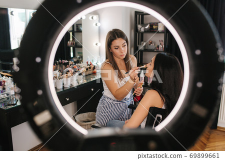Photo though the ring lamp of makeup artist making a makeup for attractive young girl. Beautiful young brunette woman 69899661