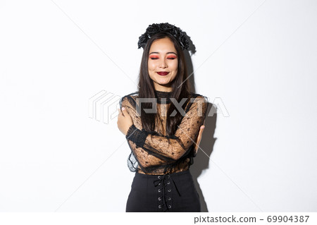 Happy cute asian woman hugging herself and smiling dreamy, wearing halloween costume of witch or widow, standing over white background 69904387