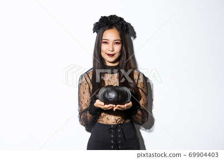 Smiling devious asian girl in witch costume, celebrating halloween, holding black pumpkin, standing over white background Smiling devious asian girl in witch costume, celebrating halloween, holding black pumpkin, standing over white background 69904403