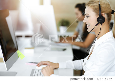 Blond business woman using headset for communication and consulting people at sunny office. Call center Blond business woman using headset for communication and consulting people at sunny office. Call center 69905776