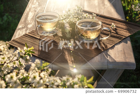 Two beautiful glass cups with green tea decorated with white cherry flowers on wooden table in spring garden in setting sun light. Two beautiful glass cups with green tea decorated with white cherry flowers on wooden table in spring garden in setting sun light. 69906732