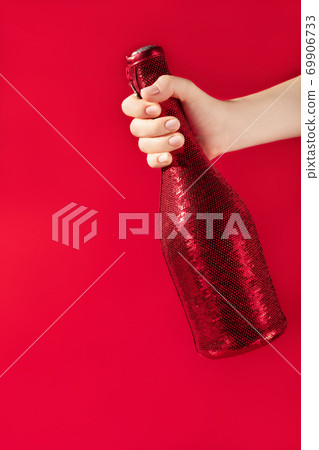 Modern design template with female hand holding champaigne bottle covered in red sequines on red background. Christmas background concept. Holiday celebration concept. Modern design template with female hand holding champaigne bottle covered in red sequines on red background. Christmas background concept. Holiday celebration concept. 69906733