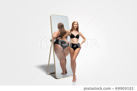 Young slim woman looking at fat girl in mirror's reflection on white background 69909181