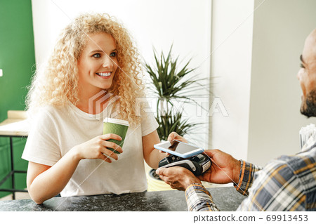 Woman customer of coffee shop paying for coffee through mobile phone using contactless technology Woman customer of coffee shop paying for coffee through mobile phone using contactless technology 69913453