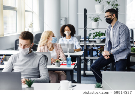 Consultation with manager about work and social distance. Millennial man and woman in protective masks talking at workplace in interior of office Consultation with manager about work and social distance. Millennial man and woman in protective masks talking at workplace in interior of office 69914557