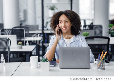 Working with modern technology and clients remotely. Friendly african american lady talking on phone with client, sitting at table with laptop in office interior Working with modern technology and clients remotely. Friendly african american lady talking on phone with client, sitting at table with laptop in office interior 69914657