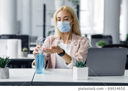 Safety and health protection at work in office. Millennial woman in protective mask with smart watch uses antiseptic at workplace with laptop for hand disinfection Safety and health protection at work in office. Millennial woman in protective mask with smart watch uses antiseptic at workplace with laptop for hand disinfection 69914658