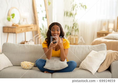 Weekend entertainments. Shocked African American woman with TV controller enjoying interesting movie indoors Weekend entertainments. Shocked African American woman with TV controller enjoying interesting movie indoors 69914659