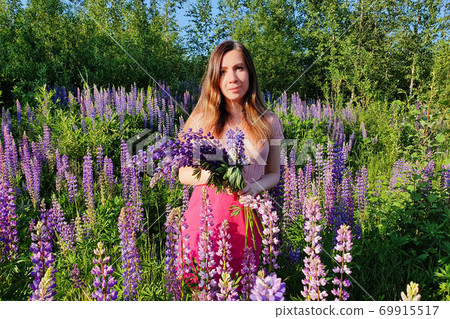 Portrait of young caucasian woman with long red hair, against the background of greenery in lupin`s field. Beautiful gathering flowers female. She is a successful florist. Portrait of young caucasian woman with long red hair, against the background of greenery in lupin`s field. Beautiful gathering flowers female. She is a successful florist. 69915517