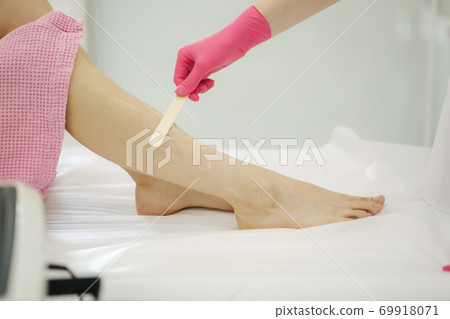 Young woman beautician in mask applies contact gel on the leg of a woman for laser hair removal in medical clinic. Close up. Pink glowes Young woman beautician in mask applies contact gel on the leg of a woman for laser hair removal in medical clinic. Close up. Pink glowes 69918071