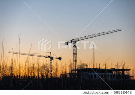 Industrial construction cranes and buildings silhouettes at a construction site on the background at sunset or dawn. Industrial construction cranes and buildings silhouettes at a construction site on the background at sunset or dawn. 69918448