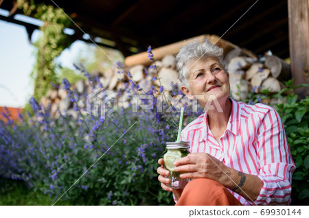 Portrait of senior woman sitting outdoors in garden, holding lemonade drink. 69930144