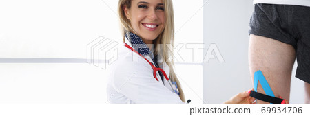 Female doctor smiles and fixes kinesio tape on male knee. Female doctor smiles and fixes kinesio tape on male knee. 69934706