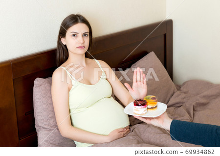 Pregnant woman lying in bed don't want to eat a cake and makes stop gesture. No sweet and dessert during pregnancy concept 69934862