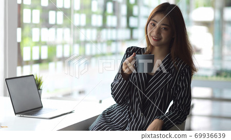Female office worker smiling and looking into camera while taking a coffee break Female office worker smiling and looking into camera while taking a coffee break 69936639