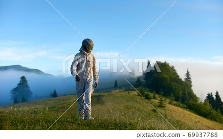 Portrait of spaceman wearing white space suit and a helmet walking alone a sunny green mountain glade 69937788