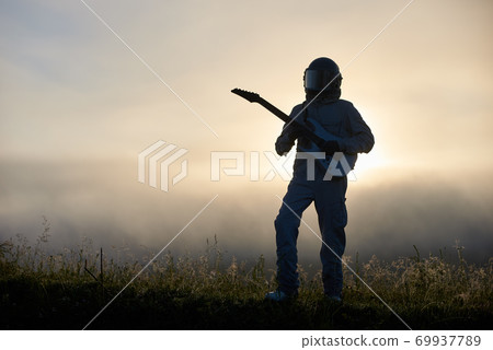 Astronaut standing on grass and playing guitar. 69937789