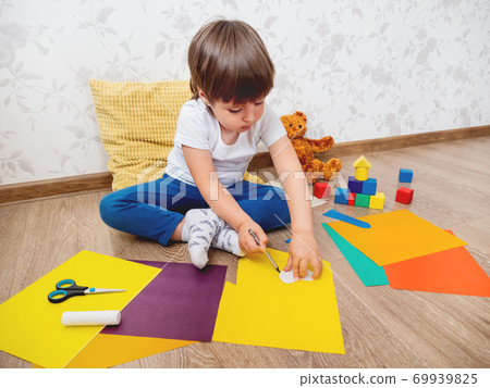 Toddler boy learns to cut colored paper with scissors. Kid sits on floor in kids room with toy blocks and teddy bear. Educational classes for children.  69939825
