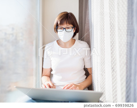 Pretty woman in medical mask works remotely from home. She sits on window sill with laptop on knees. Lockdown quarantine because of coronavirus COVID19. Self isolation at home. 69939826