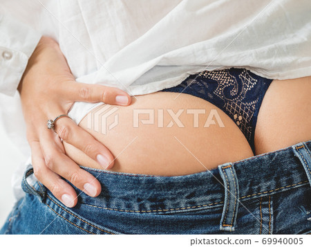 Close up photo of woman buttocks with laced thong and engagement ring on her finger. Woman in white shirt, classic blue jeans and underwear. 69940005