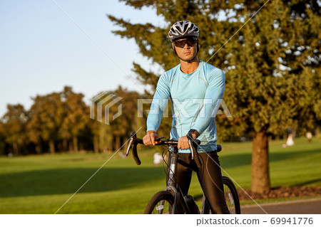 Resting after bicycle ride outdoors. Young athletic man in sportswear and protective helmet standing with his bike in park and looking at camera Resting after bicycle ride outdoors. Young athletic man in sportswear and protective helmet standing with his bike in park and looking at camera 69941776