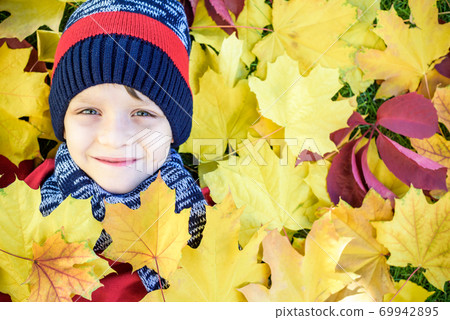 Emotional portrait of a happy and active little boy looking at the camera with a smile lying on his back on a carpet of yellow fallen leaves in the autumn park. Positive emotions. Emotional portrait of a happy and active little boy looking at the camera with a smile lying on his back on a carpet of yellow fallen leaves in the autumn park. Positive emotions. 69942895