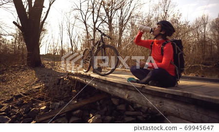 Happy woman sits wooden bridge over river, listening music in headphones with smartphone hands and drinks water white bottle. cyclist listens song in nature. athlete near bike hand on plastic drink 69945647
