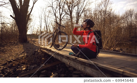 Happy woman sits wooden bridge over river, listening music in headphones with smartphone hands and drinks water white bottle. cyclist listens song in nature. athlete near bike hand on plastic drink Happy woman sits wooden bridge over river, listening music in headphones with smartphone hands and drinks water white bottle. cyclist listens song in nature. athlete near bike hand on plastic drink 69945648