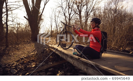 Happy caucasian woman cyclist makes a photo of herself a selfie on the phone while sitting on the bridge over a river in the forest in sunny weather 69945649