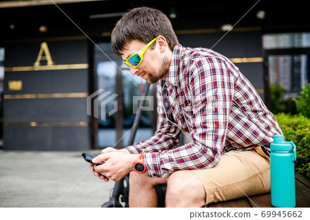 Modern ecological clean transport theme. A man resting on a bench and holding a smart phone in his hand after riding an eco electric kick scooter in the city. Male riding a scooter checking messages 69945662