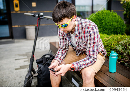 Caucasian man uses modern technologies Internet and cellular communication on smartphone while sitting on bench in city near electric scooter. Ecology and environmentally friendly urban transport 69945664