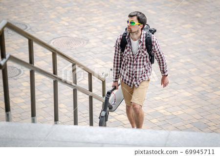 Man walking with folded electric scooter up stairs in city near modern building. Ecological technological lifestyle. E-Mobility. Person Carrying Electric Scooter Folded Position. Eco urban transport Man walking with folded electric scooter up stairs in city near modern building. Ecological technological lifestyle. E-Mobility. Person Carrying Electric Scooter Folded Position. Eco urban transport 69945711